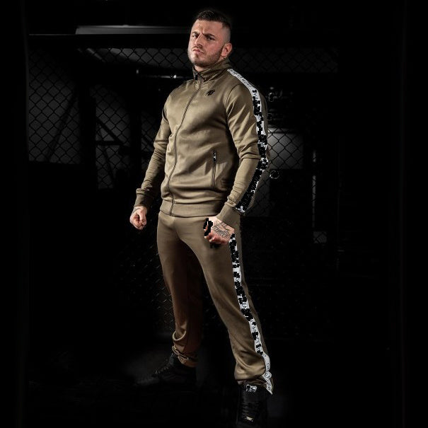 Wellington Track Jacket takki värissä khaki. Kuvattu mallin päällä. Logo vasemmalla. Valkoiset raidat mustalla tekstillä hihoissa.